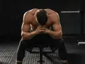 Uomo muscoloso seduto in palestra, testa china, mani giunte. Potrebbe essere un momento di riflessione dopo un duro allenamento o un **test sovrallenamento**.