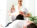 Un fisioterapista esegue un trattamento alla gamba di un paziente, con un poster anatomico sullo sfondo.