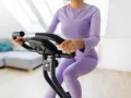 Donna in tuta viola pedala su cyclette, riabilitazione post protesi ginocchio.