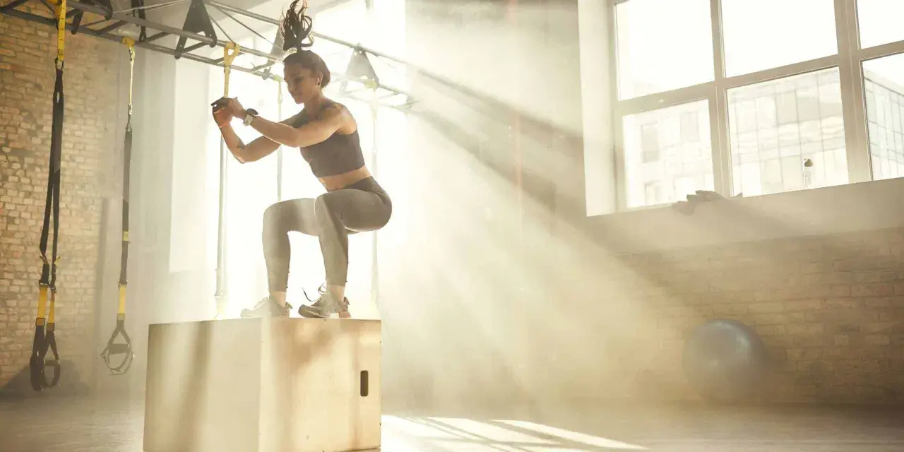 Donna esegue un box jump, dimostrando diversi tipi di forza in palestra, con raggi di sole che filtrano.