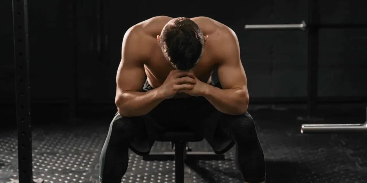 Uomo muscoloso seduto in palestra, testa china, mani giunte. Potrebbe essere un momento di riflessione dopo un duro allenamento o un **test sovrallenamento**.