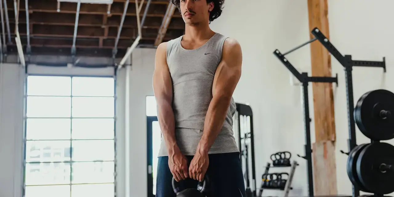Giovane uomo in palestra, con kettlebell in mano, mostra i suoi muscoli lombari e braccia scolpite.