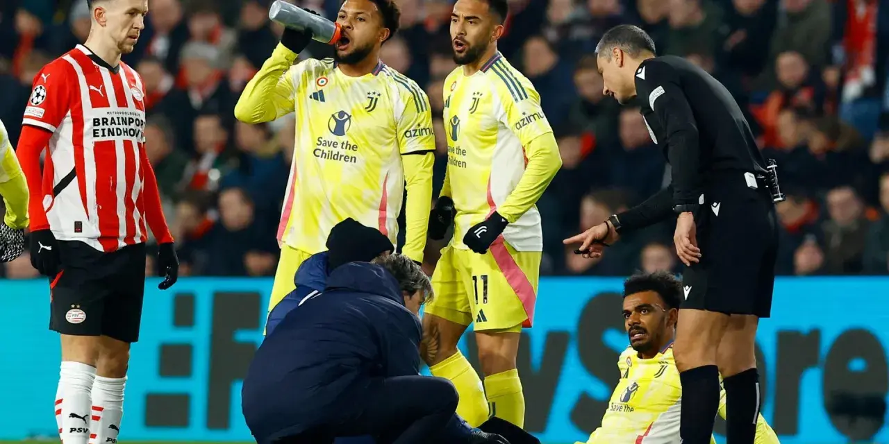 Giocatori di calcio in campo, uno è a terra, assistito dallo staff medico. L'arbitro osserva attentamente, preoccupato per i possibili infortuni nel calcio.