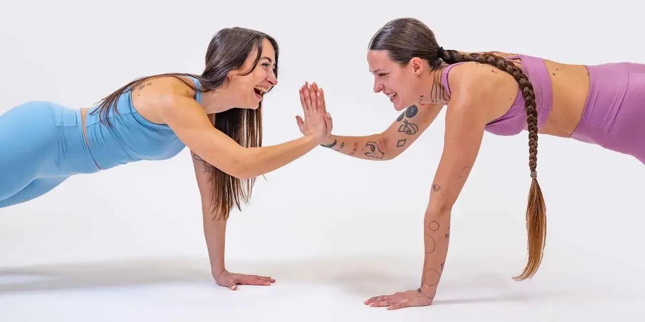Due donne in posizione di plank si danno il cinque, un allenamento post parto che rafforza il corpo e lo spirito.