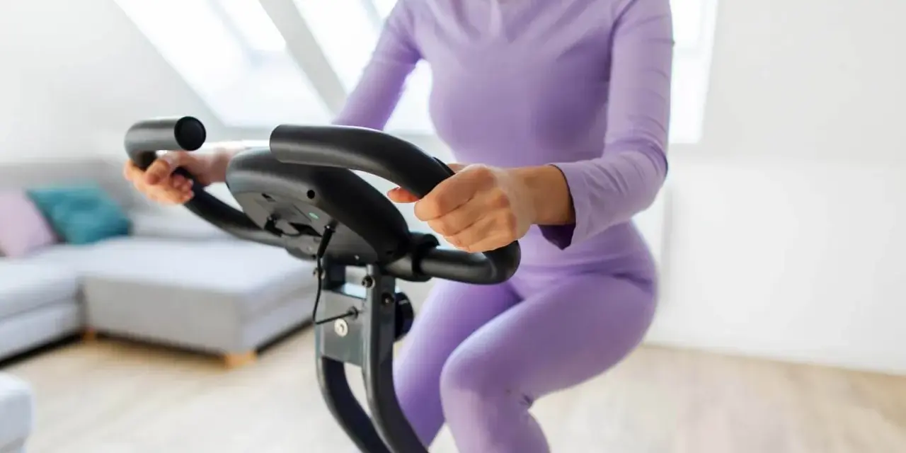 Donna in tuta viola pedala su cyclette, riabilitazione post protesi ginocchio.