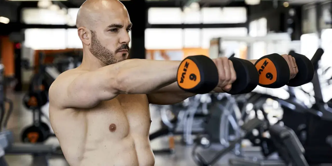 Uomo calvo in palestra esegue alzate laterali con manubri da 6kg. Allenamento 3x10+10 per spalle definite.