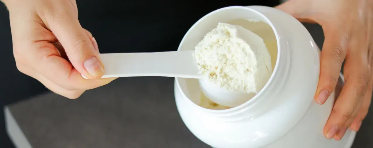 Mano che preleva proteine in polvere da un barattolo bianco con un misurino. Ideale per il recupero post-allenamento.