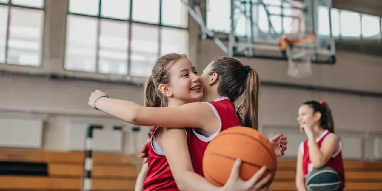 Due giocatrici di basket si abbracciano, una tiene un pallone. Forse si allenano con esercizi mobilità anche e bacino.