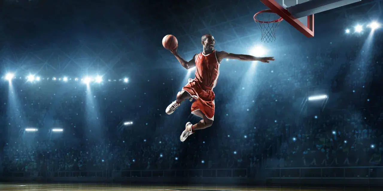 Un giocatore di basket in aria, con la palla in mano, dimostra la sua forza muscolare con un salto spettacolare sotto le luci dell'arena.