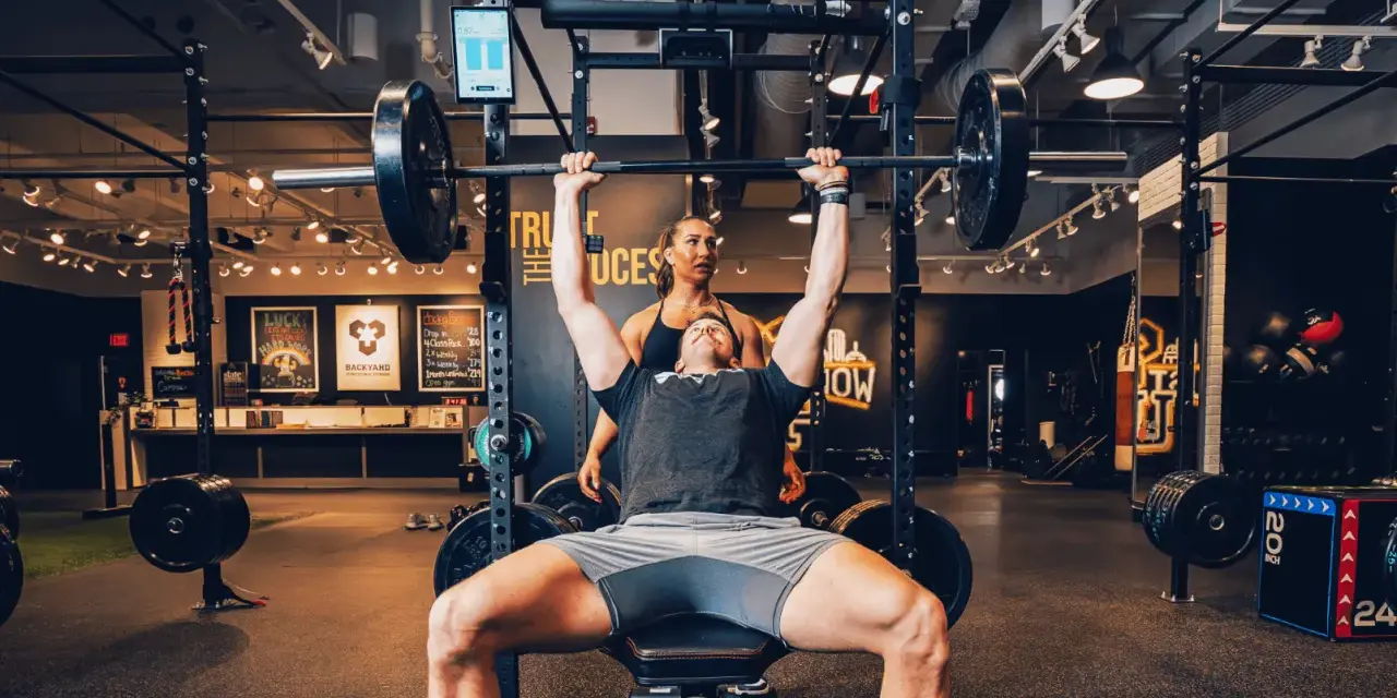 Uomo solleva bilanciere con donna che lo osserva, allenamento che stimola diversi tipi di fibre muscolari.