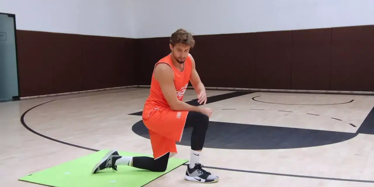 Atleta in ginocchio su un tappetino verde, esegue un esercizio di riscaldamento caviglie in palestra.