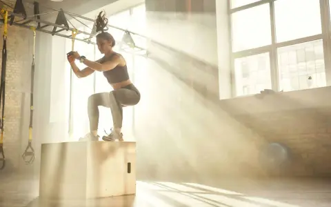Donna esegue un box jump, dimostrando diversi tipi di forza in palestra, con raggi di sole che filtrano.