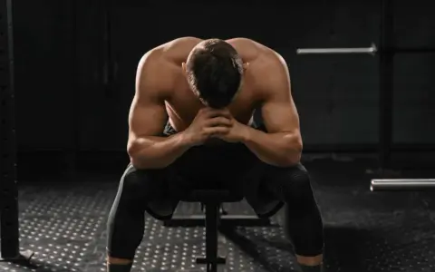 Uomo muscoloso seduto in palestra, testa china, mani giunte. Potrebbe essere un momento di riflessione dopo un duro allenamento o un **test sovrallenamento**.