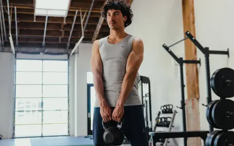 Giovane uomo in palestra, con kettlebell in mano, mostra i suoi muscoli lombari e braccia scolpite.