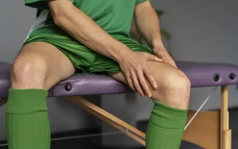 Atleta con ginocchio lasso, seduto su lettino da massaggio, con mani che stringono la gamba.