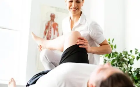 Un fisioterapista esegue un trattamento alla gamba di un paziente, con un poster anatomico sullo sfondo.