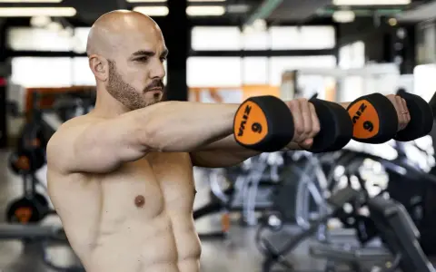 Uomo calvo in palestra esegue alzate laterali con manubri da 6kg. Allenamento 3x10+10 per spalle definite.