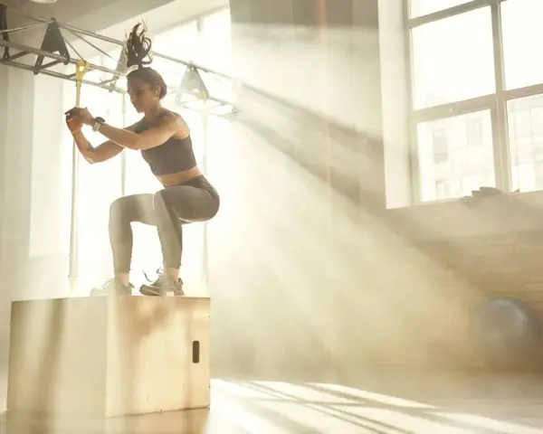 Donna esegue un box jump, dimostrando diversi tipi di forza in palestra, con raggi di sole che filtrano.