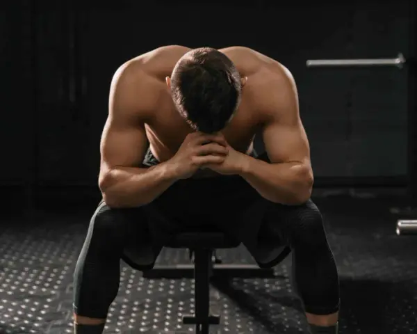 Uomo muscoloso seduto in palestra, testa china, mani giunte. Potrebbe essere un momento di riflessione dopo un duro allenamento o un **test sovrallenamento**.