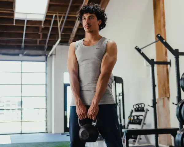 Giovane uomo in palestra, con kettlebell in mano, mostra i suoi muscoli lombari e braccia scolpite.