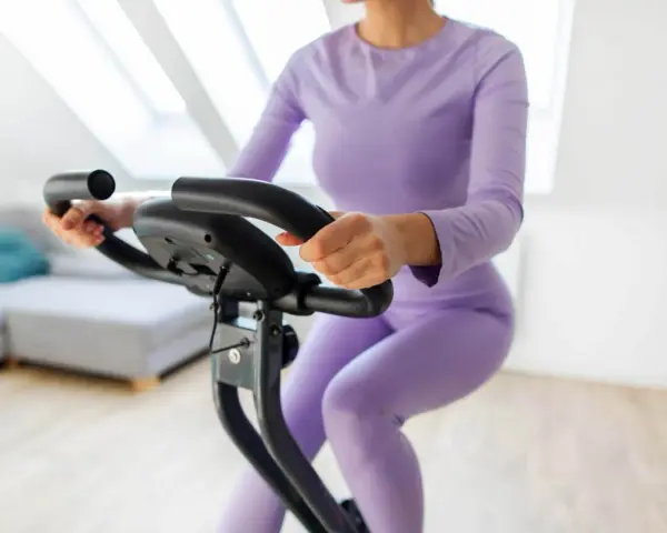 Donna in tuta viola pedala su cyclette, riabilitazione post protesi ginocchio.