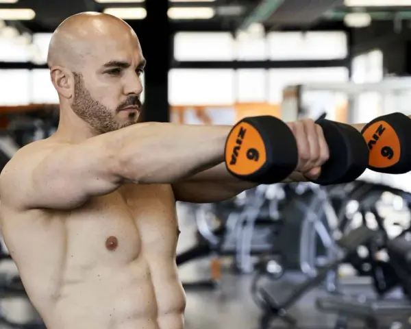 Uomo calvo in palestra esegue alzate laterali con manubri da 6kg. Allenamento 3x10+10 per spalle definite.