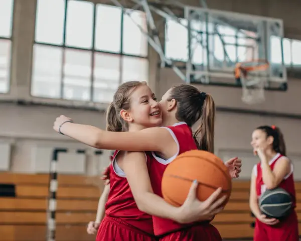 Due giocatrici di basket si abbracciano, una tiene un pallone. Forse si allenano con esercizi mobilità anche e bacino.