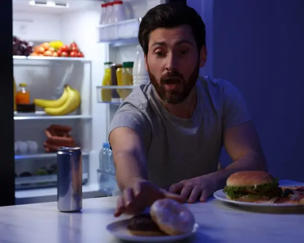 Uomo con espressione sorpresa allunga la mano verso ciambelle e pizza, con il frigorifero aperto alle sue spalle, una tentazione notturna che sfida la finestra metabolica.