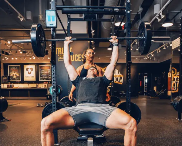 Uomo solleva bilanciere con donna che lo osserva, allenamento che stimola diversi tipi di fibre muscolari.