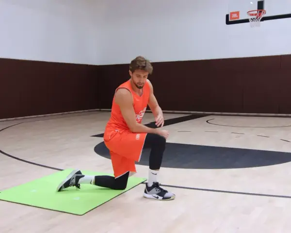 Atleta in ginocchio su un tappetino verde, esegue un esercizio di riscaldamento caviglie in palestra.