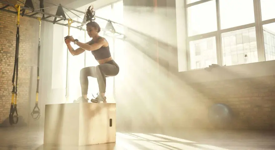 Donna esegue un box jump, dimostrando diversi tipi di forza in palestra, con raggi di sole che filtrano.