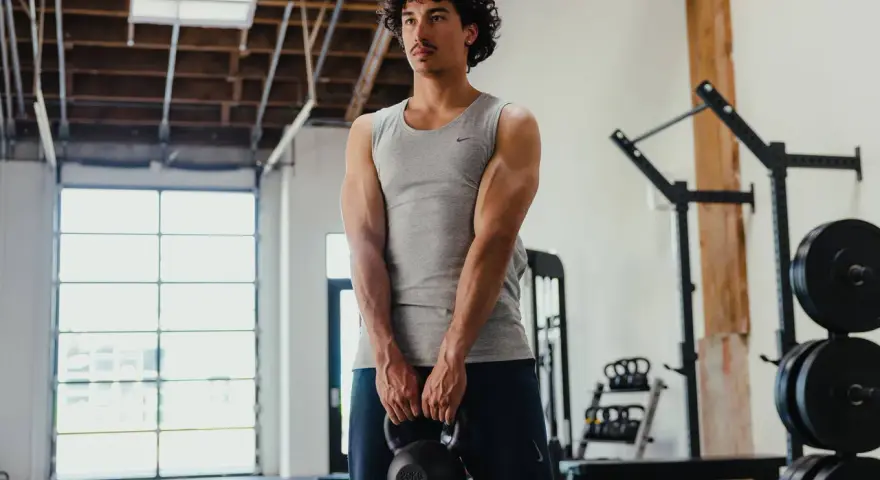 Giovane uomo in palestra, con kettlebell in mano, mostra i suoi muscoli lombari e braccia scolpite.