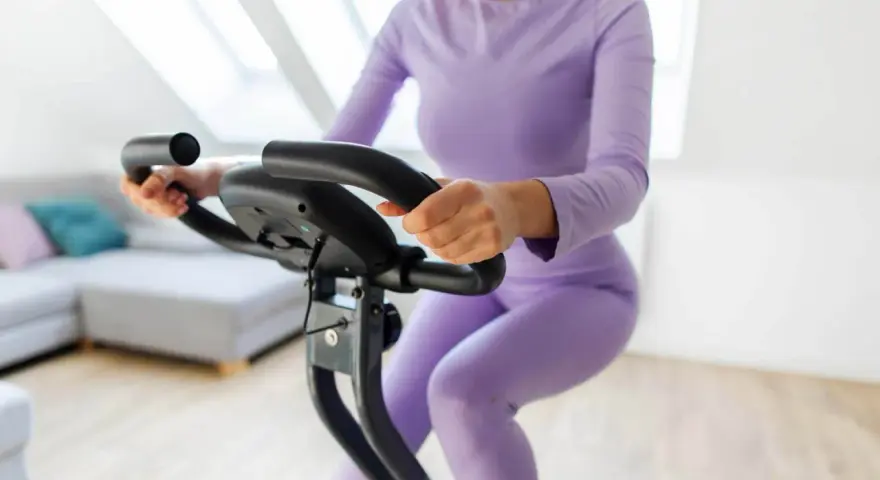 Donna in tuta viola pedala su cyclette, riabilitazione post protesi ginocchio.