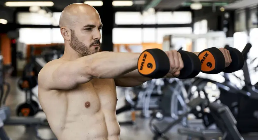 Uomo calvo in palestra esegue alzate laterali con manubri da 6kg. Allenamento 3x10+10 per spalle definite.