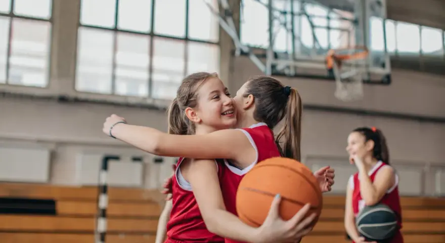 Due giocatrici di basket si abbracciano, una tiene un pallone. Forse si allenano con esercizi mobilità anche e bacino.