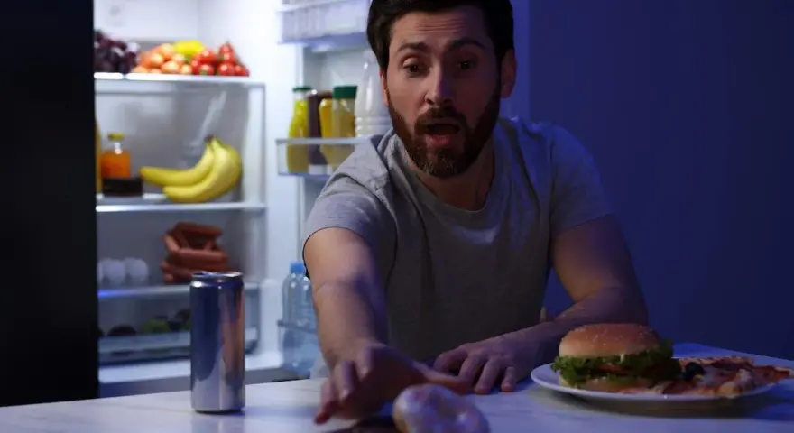 Uomo con espressione sorpresa allunga la mano verso ciambelle e pizza, con il frigorifero aperto alle sue spalle, una tentazione notturna che sfida la finestra metabolica.
