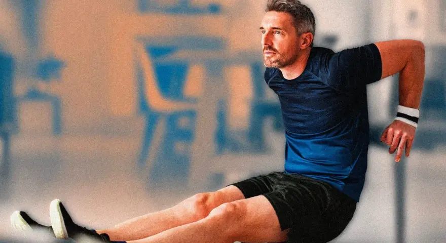 Uomo con polsino bianco esegue allenamento con sedia, piegando le braccia. Gambe tese in avanti, scarpe sportive.