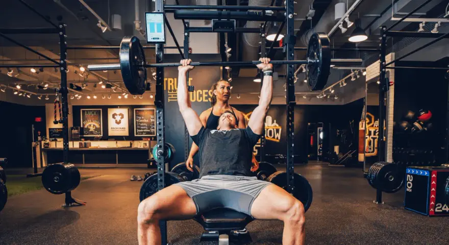 Uomo solleva bilanciere con donna che lo osserva, allenamento che stimola diversi tipi di fibre muscolari.