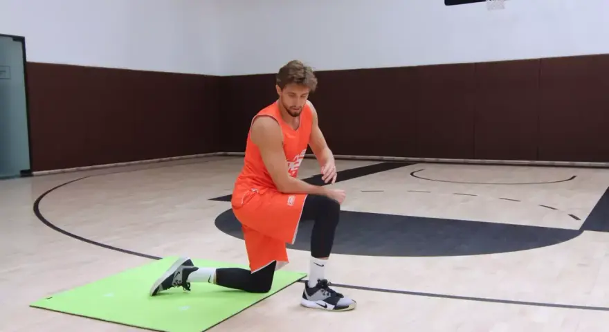 Atleta in ginocchio su un tappetino verde, esegue un esercizio di riscaldamento caviglie in palestra.