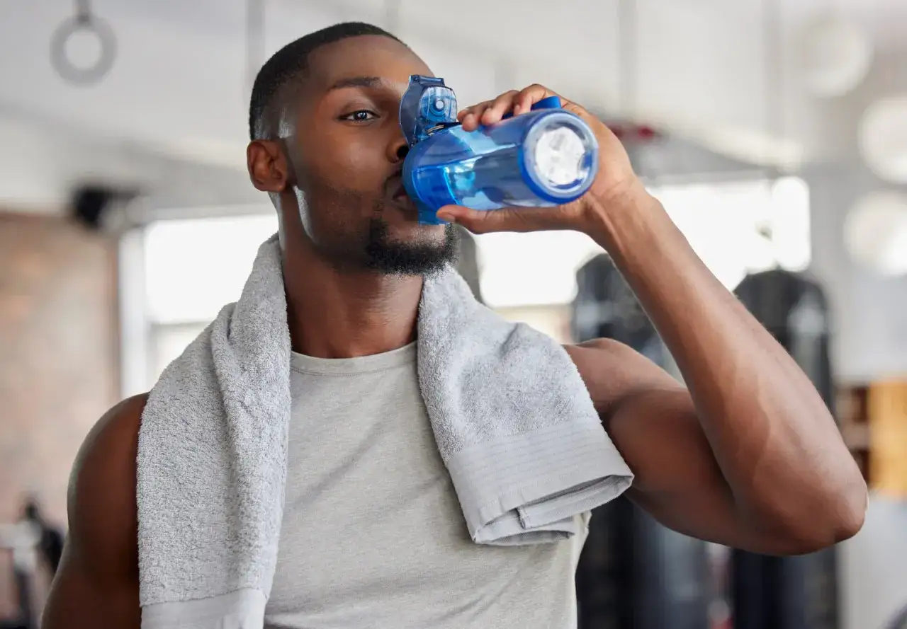 Uomo con asciugamano in palestra beve da borraccia blu. L'acqua è la scelta migliore per cosa bere durante l'allenamento in palestra.