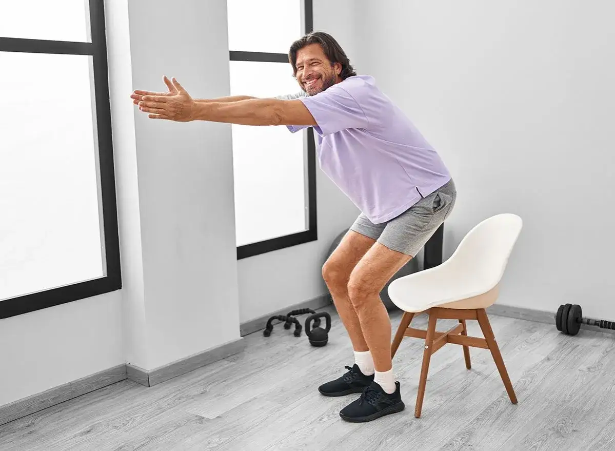 Uomo sorridente fa squat assistito da una sedia, un allenamento con sedia efficace per gambe e glutei.