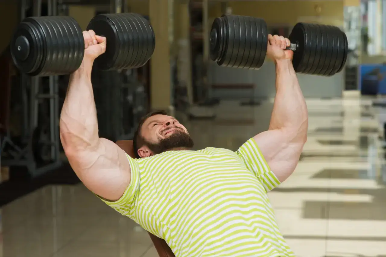 Uomo con barba solleva manubri pesanti in palestra. Si allena con 3x10+10, concentrato e con muscoli tesi.