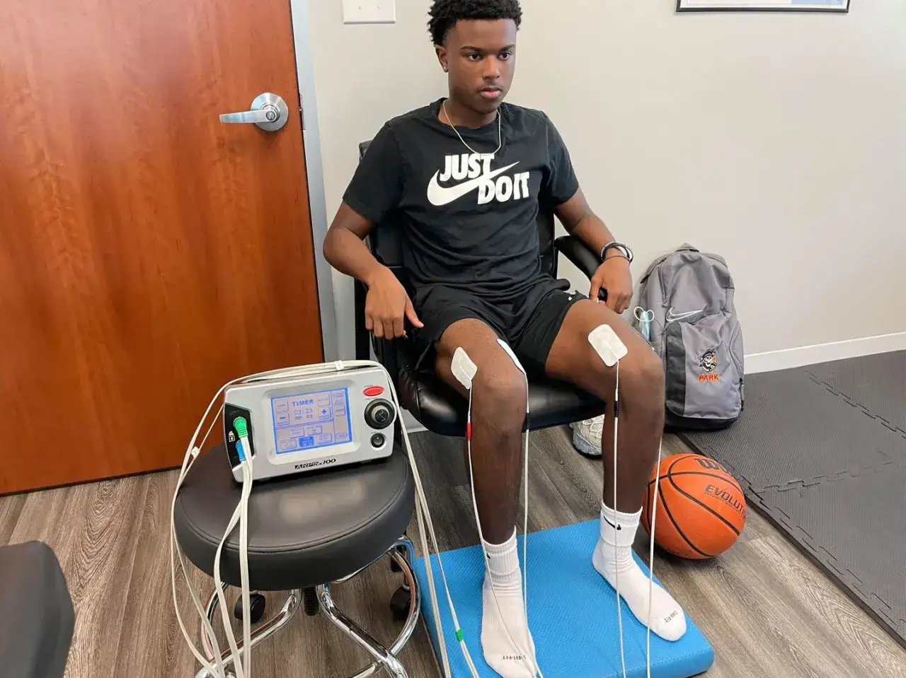 Giovane atleta con elettrodi sulle ginocchia per un trattamento, forse per un ginocchio lasso, seduto vicino a un dispositivo medico e un pallone da basket.