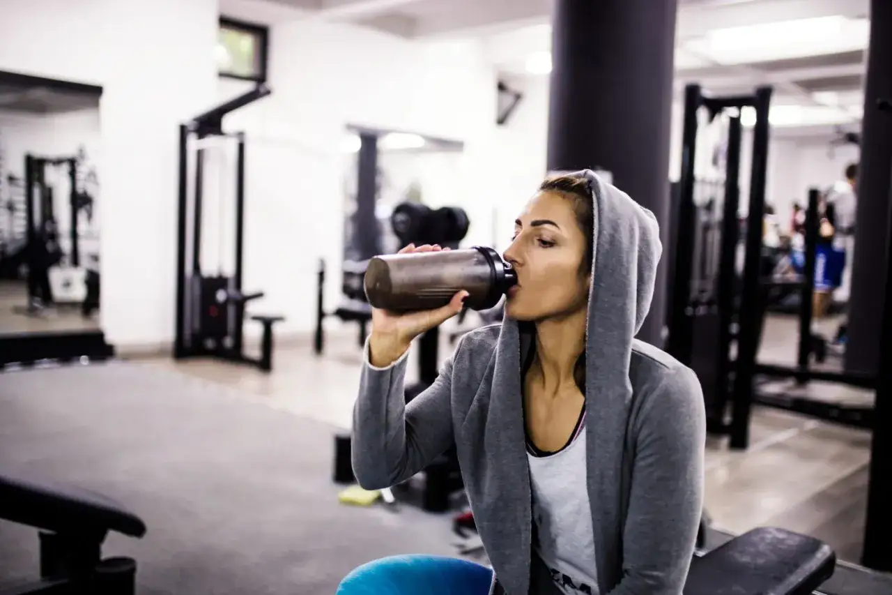 Donna con cappuccio beve da uno shaker, integrando proteine in polvere prima o dopo allenamento in palestra.