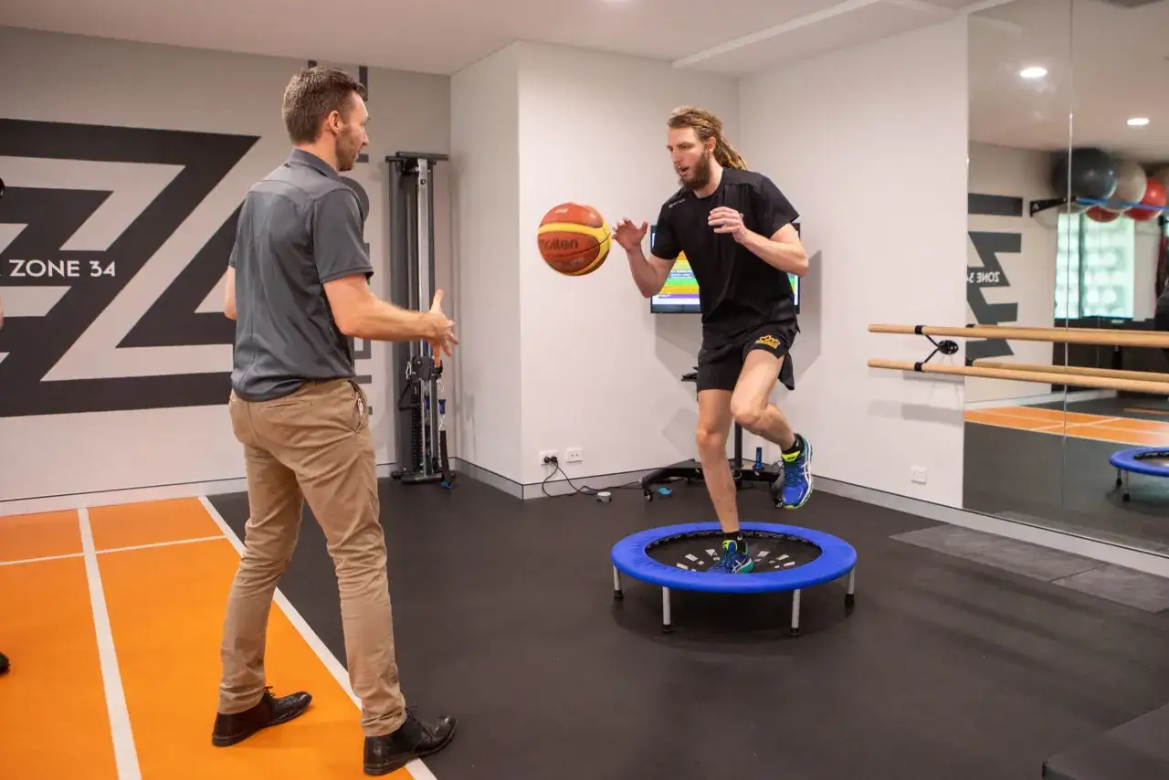 Fisioterapista osserva paziente con infortunio caviglia che si allena su trampolino con palla da basket.